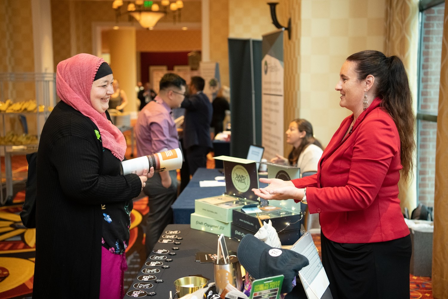 Two women engage in conversation at an event booth, surrounded by promotional items, while other attendees and booths are visible in the background.