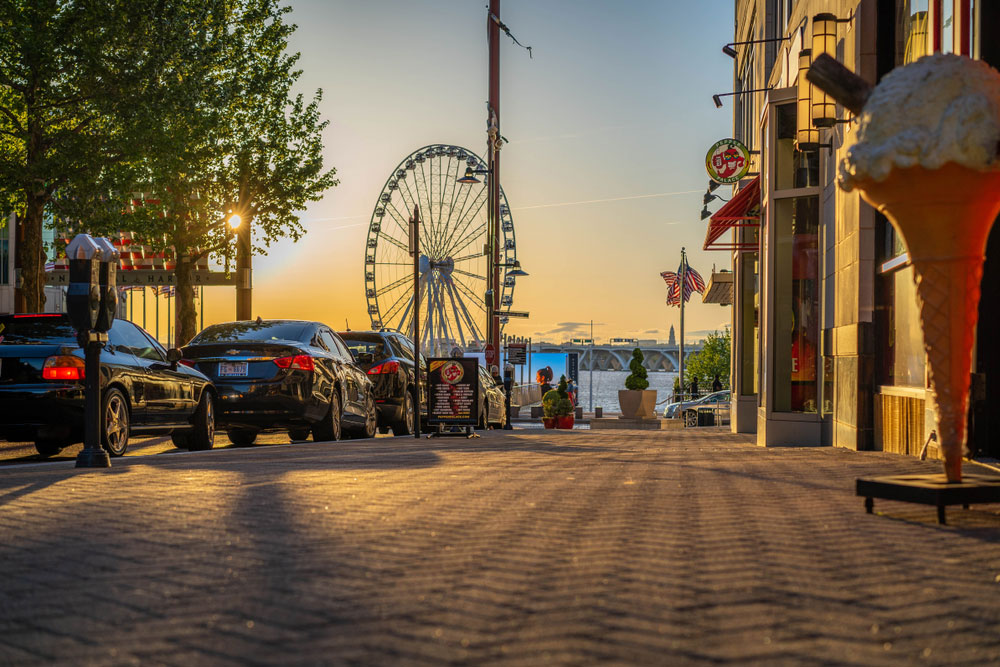 National harbor street lined with cars and shops with a ferris wheel and ocean in the background