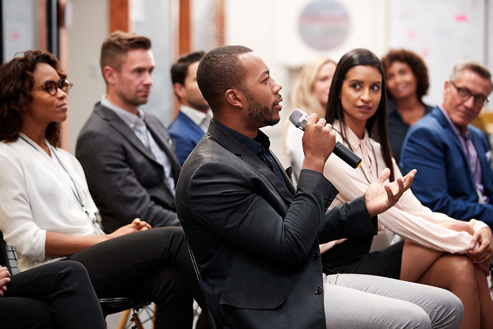 A man in a suit speaks into a microphone during a group meeting or seminar, surrounded by seated people listening attentively.