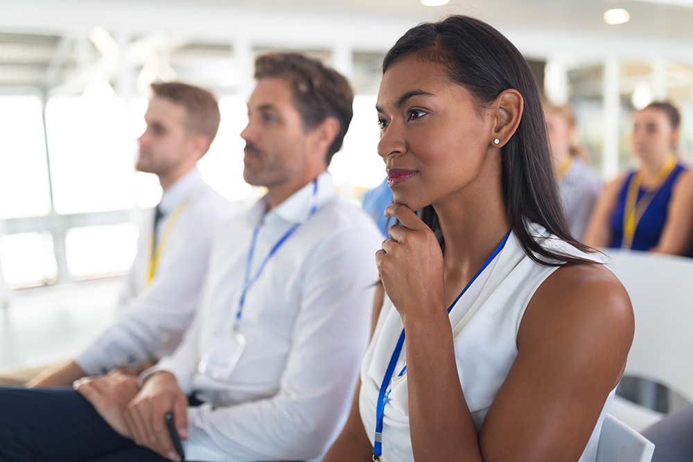 People seated in a conference room, attentively listening, wearing lanyards. The focus is on a woman in the foreground with a thoughtful expression.