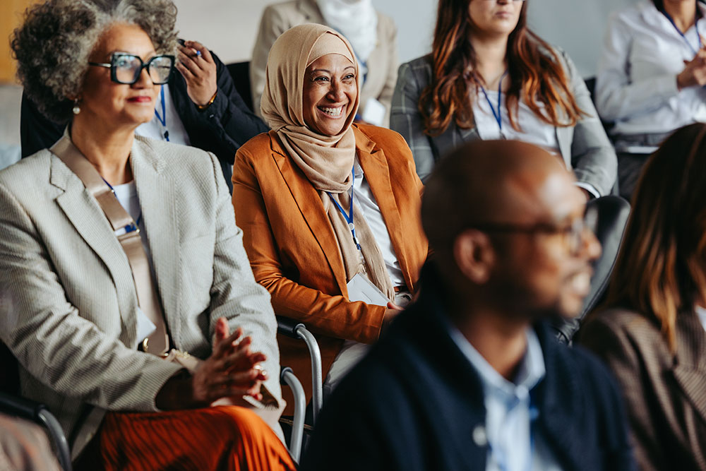 A diverse group of people sitting and smiling in a conference setting. The focus is on a woman smiling wearing a hijab.