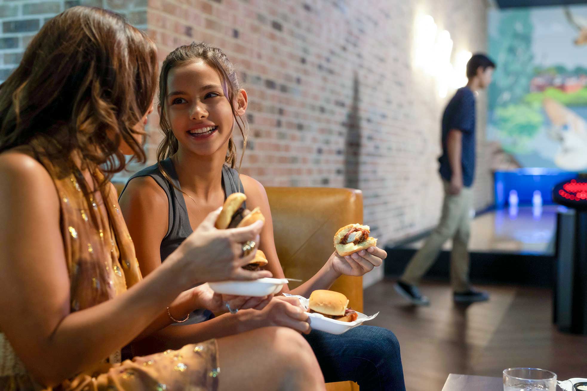 Two women sharing a meal at a bowling alley, with bowling lanes and colorful decor in the background.