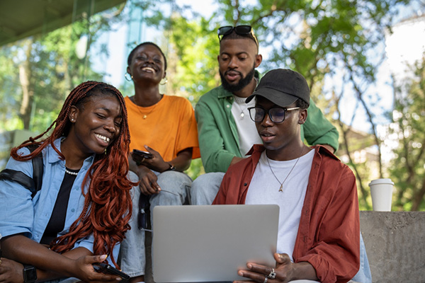 A group of four young people on a bench, focused on a laptop, enjoying a collaborative moment in a park setting.